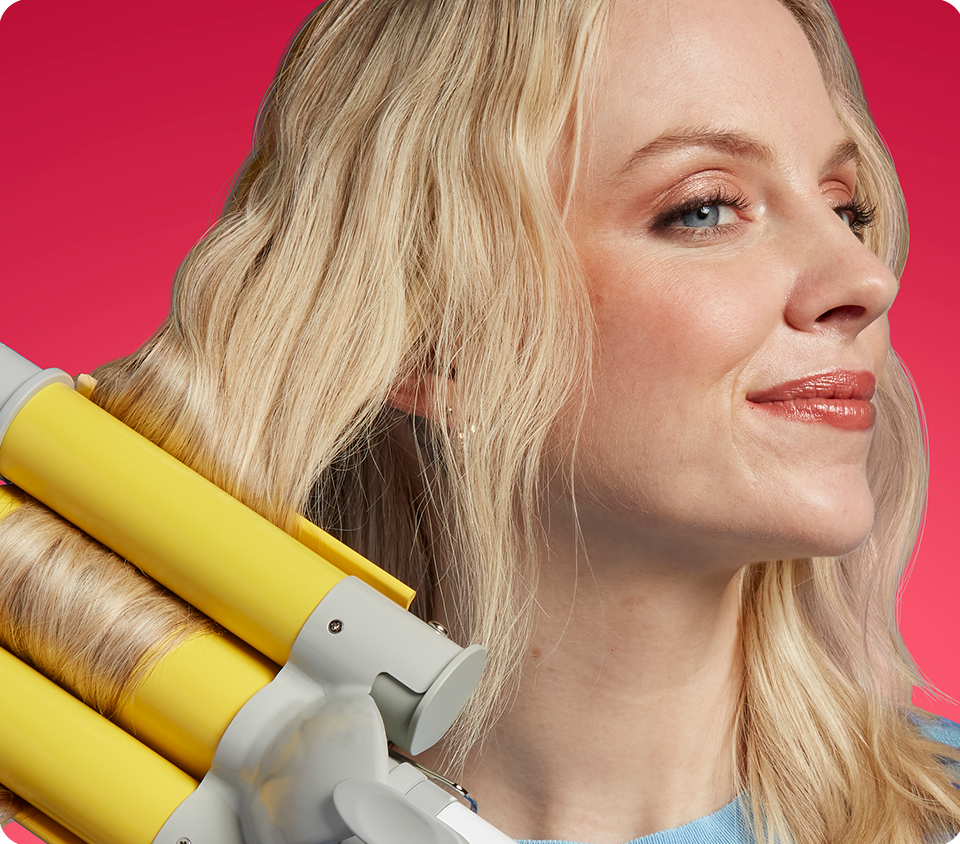 Woman using Conair Style Chemistry triple-barrel waver attachment on long straight hair, against red background
