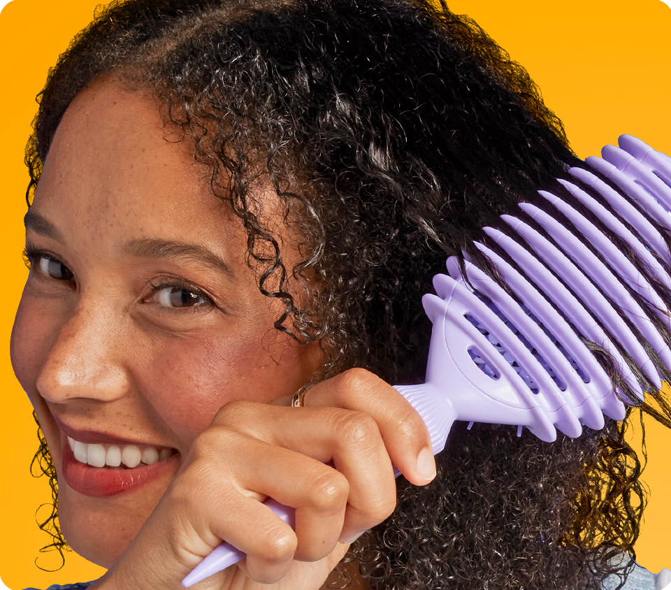 Woman brushing through natural curls with Conair Curl Define Pro hairbrush, against yellow background 